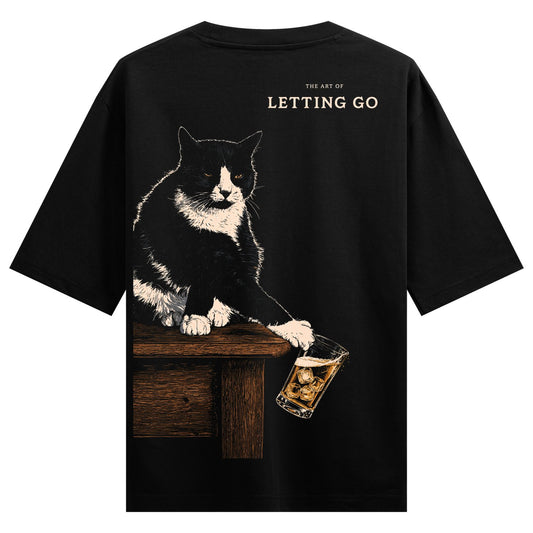 Cat Letting Go Oversized T-Shirt — a black and white cat sits on a wooden table holding a whiskey glass with ice cubes, text