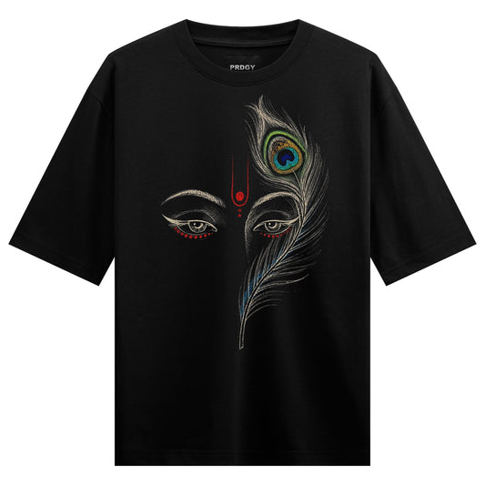 A black t-shirt displays a split face with detailed eyes and a vibrant peacock feather on one side featuring a red tilak a...