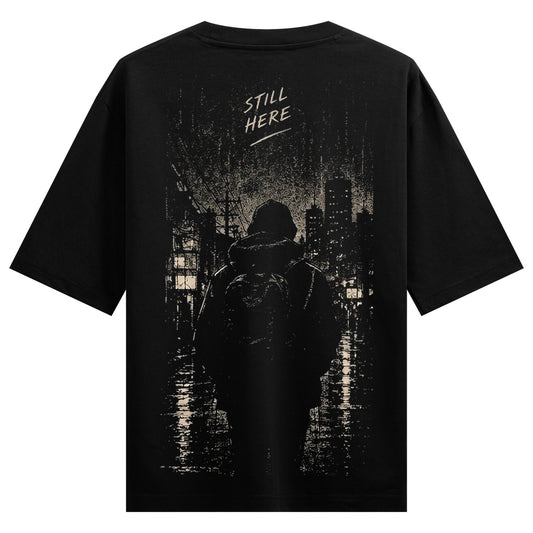 Urban Shadows Oversized T-Shirt — a hooded figure with a backpack stands in the rain, facing a city skyline under a dark sky